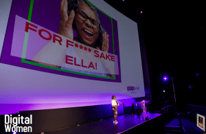 Ella is speaking on stage with a power point slide behind her.